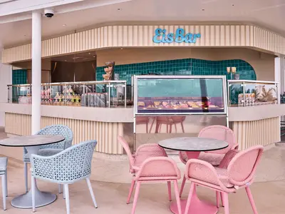 Die helle Eis Bar an Bord der Mein Schiff Relax mit Pastellfarben. Eine Eisvitrine, dahinter türkise Fliesen und eine Theke mit Sitzmöglichkeiten in Rosa und Hellblau sind zu sehen.