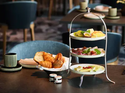 Eine Etagere mit frischem Obst, Aufschnitt und Käse steht auf einem Tisch in einem Restaurant der Mein Schiff Relax. Daneben ein Korb mit Gebäck, zwei Marmeladengläser und eine Tasse.