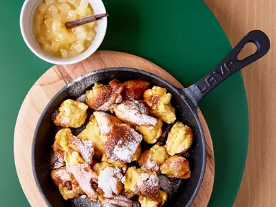 Eine gusseiserne Pfanne gefüllt mit lockerem, zerrissenem Kaiserschmarrn, reichlich mit Puderzucker bestreut, daneben eine Schale mit Apfelkompott, serviert auf einem Holzbrett an Bord der Mein Schiff Relax.