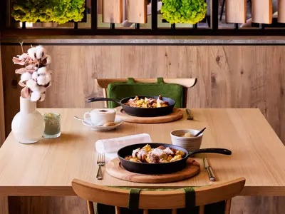 Blick auf einen gedeckten Holztisch im Restaurant der Mein Schiff Relax. Zwei gusseiserne Pfannen mit Kaiserschmarrn und Puderzucker, dazu Kaffee und Apfelkompott. Eine Vase mit Baumwollzweigen steht links.