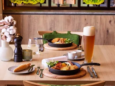 Ein schön gedeckter Holztisch mit zwei gusseisernen Pfannen mit Rösti, Spiegelei, Bacon und Rucola an Bord der Mein Schiff Relax. Dazu ein großes Glas Bier und ein Brötchen.