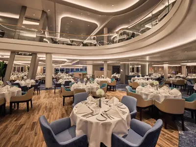 Umfassender Blick in das moderne, elegant eingerichtete 'Atlantik' Restaurant an Bord der Mein Schiff Relax, komplett mit weiß gedeckten Tischen und bequemen Stühlen in Blau- und Beigetönen auf zwei Ebenen.