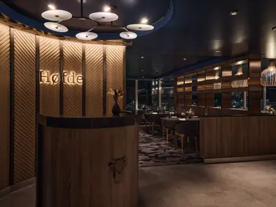 Eingangsbereich des Restaurants Høfde auf der Mein Schiff Relax. Holzwände mit Diagonalmuster, beleuchtetes Logo. Tische und Fenster mit Nachtblick sind im Hintergrund zu sehen.