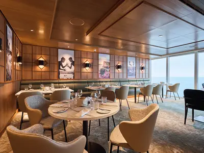 Ein modernes Restaurant an Bord der Mein Schiff Relax mit Holzwänden, stilvollen Leuchten, gedeckten Tischen und großen Fenstern mit Blick auf das Meer. Es sind keine Personen zu sehen.