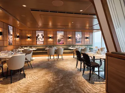 Modernes Restaurant an Bord der Mein Schiff Relax. Helle Tische, bequeme Stühle und eine lange Bank. Holzwände mit Kunst und warmem Licht. Elegant und einladend.