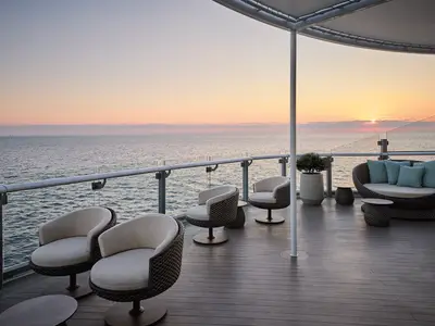 Ausblick auf das Meer bei Sonnenuntergang von einer modernen Terrasse an Bord der Mein Schiff Relax. Elegante Rattan-Loungemöbel stehen auf dem Holzdeck bereit.
