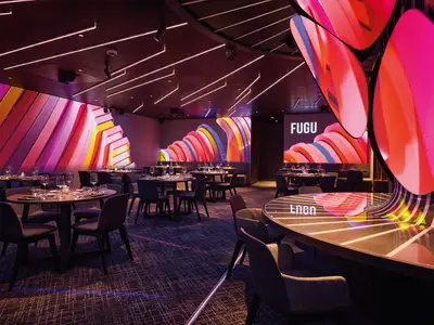 Blick in das moderne Spezialitätenrestaurant FUGU an Bord der Mein Schiff Relax. Gedeckte Tische stehen vor Wänden mit farbiger, abstrakter Digitalprojektion und pinkfarbenen Lichtinstallationen an Decke und Säulen.