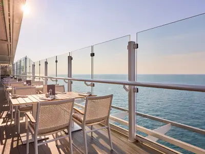 Ein heller Außenbereich des X-View Restaurants an Bord der Mein Schiff Relax mit Holztischen und geflochtenen Stühlen. Im Hintergrund das weite Meer unter klarem Himmel.