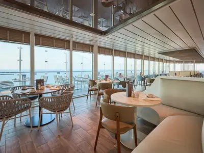 Der helle, moderne X-View Restaurantbereich an Bord der Mein Schiff Relax bietet grosse Fenster mit Blick auf das Meer. Mehrere Tische mit Stühlen und bequemen Bänken sind elegant gedeckt.