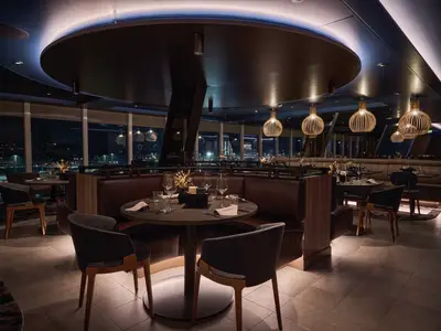 Restaurant Hoefde an Bord der Mein Schiff Relax bei Nacht. Elegante dunkle Tische und Lederbänke. Große Fenster mit Blick auf beleuchtete Skyline. Moderne Hängelampen schaffen eine warme Atmosphäre.