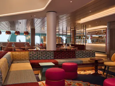 Das moderne Mein Schiff Bistro an Bord der Mein Schiff Relax zeigt farbenfrohe Sitzbereiche, darunter eine Patchwork-Couch. Im Hintergrund ist die hell beleuchtete Theke sichtbar. Ohne Personen.