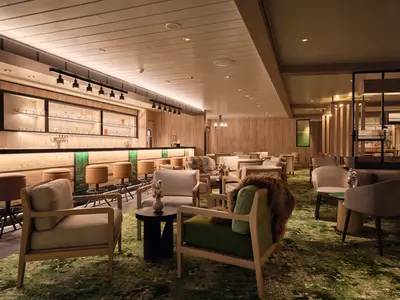 Elegantes Chalet Restaurant und Bar an Bord der Mein Schiff Relax. Holzwände, grüne Bar mit Hockern, helle Sessel und Tische mit Fellen. Beleuchtete Regale mit Flaschen.