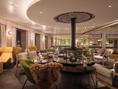 Blick in die elegante Chalet-Lounge der Mein Schiff Relax mit zentralem, verglastem Kamin und vielfältigen Sitzbereichen in warmen Farben und Holztönen. Große Fenster und indirekte Beleuchtung sorgen für eine gemütliche Atmosphäre.