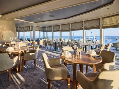 Ein helles, modernes Restaurant an Bord der Mein Schiff Relax mit vielen Tischen, die zum Essen einladen. Grosse Fenster bieten einen atemberaubenden Blick auf das weite Meer und den Aussenbereich. Die Tische sind elegant gedeckt, die Stühle bequem und in Naturtönen gehalten.
