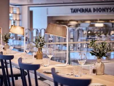 Ein stilvolles Restaurant an Bord der Mein Schiff Relax, die Taverna Dionysos. Mehrere elegant gedeckte Holztische mit modernen Lampen, Gläsern und olivengrünen Pflanzenvasen stehen bereit. Im Hintergrund ist der Schriftzug des Restaurants zu sehen.