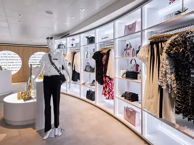 Eine stilvolle Modeboutique an Bord der Mein Schiff Relax. Helle Regale mit LED-Beleuchtung zeigen Designerkleidung und Handtaschen. Im Vordergrund steht eine Schaufensterpuppe. Links sind runde Holzdisplays mit Brillen zu sehen.