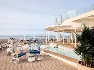 Ein modernes Sonnendeck an Bord der Mein Schiff Relax mit bequemen Loungemöbeln, einem flachen Pool und zahlreichen Liegen. Im Hintergrund ist das weite Meer sichtbar.