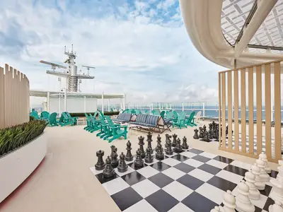 Ein weitläufiges Sonnendeck der Mein Schiff Relax mit tealfarbenen Stühlen, Lounge-Sofas und einem großen Schachfeld. Im Hintergrund sind das Meer und der Schiffsmast zu sehen.