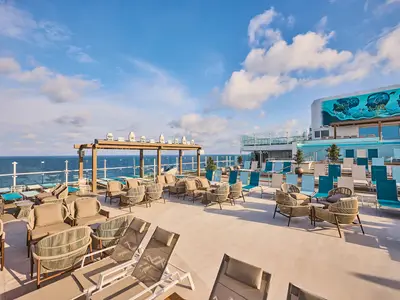 Sonnendeck der Mein Schiff Relax mit Lounge-Sitzgruppen und Liegestühlen in Blau und Beige. Pool, maritime Wandmalerei und Meer unter blauem Himmel im Hintergrund.
