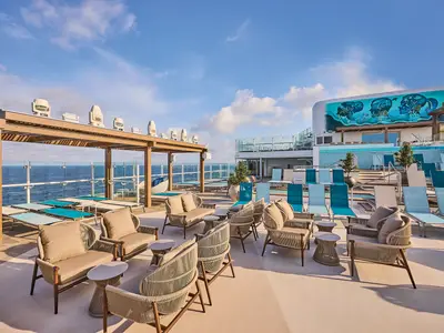 Sonnendeck der Mein Schiff Relax mit modernen Lounge-Möbeln, Liegestühlen am Pool, einer 'Sea La Vie'-Wandmalerei und weitem Meerblick unter blauem Himmel.