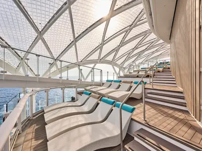 Ein terrassiertes Sonnendeck mit weissen Liegen und blauen Kopfkissen unter einem Glasdach. Blick auf das Meer, an Bord der Mein Schiff Relax.