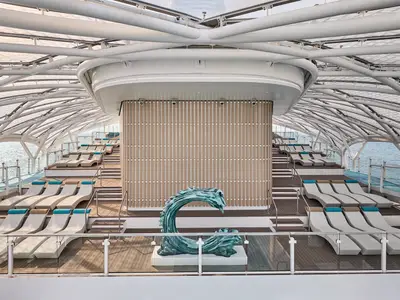Sonnendeck der Mein Schiff Relax mit modernen Liegestühlen unter geschwungenem Dach. Türkise Wellen-Skulptur mittig. Meerblick.