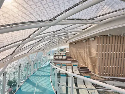Panoramablick auf die teilweise überdachte Jogging-Strecke mit Liegestühlen und Glasgeländer an Bord der Mein Schiff Relax. Das Meer ist seitlich sichtbar.