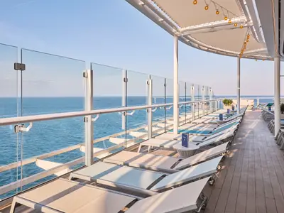 Sonniges X-Sonnendeck der Mein Schiff Relax. Moderne Liegestühle. Glasbrüstung mit Blick auf weites, blaues Meer. Sonnenschutzdach mit Lichterketten.
