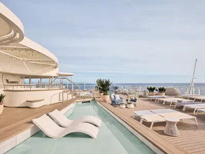 Das X-Sundeck der Mein Schiff Relax. Flacher Pool mit weißen Liegen. Holzdeck mit modernen Sonnenliegen, Sesseln und einer Bar. Weitblick auf das Meer und den Horizont.