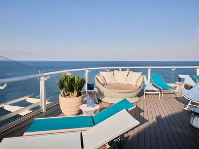 Sonniges X-Sonnendeck der Mein Schiff Relax mit Meerblick. Auf dem Holzdeck stehen Liegestühle und eine runde Lounge. Eine Glasreling gewährt ungestörte Aussicht auf das weite Meer.