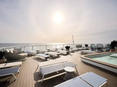 Das sonnige Sonnendeck der Mein Schiff Relax mit Liegen, Lounge-Moebeln und einem Pool. Weitblick auf das offene Meer unter hellem Himmel.