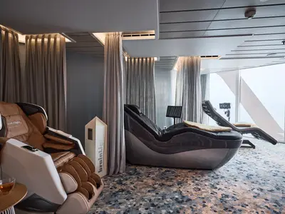 Blick in die helle Spa-Lounge der Mein Schiff Relax mit modernen Massage- und Relaxliegen. Grosse Fenster bieten Meerblick. Ein gemusterter Teppich und indirekte Beleuchtung dominieren den Raum.