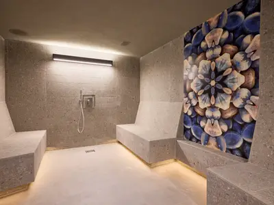 Moderne Dampfsauna an Bord der Mein Schiff Relax. Graue Fliesen, beleuchtete Sitzbänke, eine Kunstwand mit blauem Kieselsteinmuster und eine Dusche.