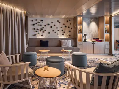 Ein moderner, gemuetlicher Sitzbereich an Bord der Mein Schiff Relax. Helle Vorhaenge, eine lange Couch mit Kissen, runde Hocker und Holzsessel auf einem gemusterten Teppich. Im Hintergrund eine Teestation und eine Wanddekoration mit sternfoermigen Elementen.