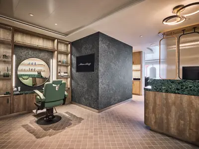 Modern eingerichteter Friseursalon an Bord der Mein Schiff Relax. Grüner Friseursessel vor Spiegel. Holzregale mit Produkten, Theke mit grünem Terrazzo.