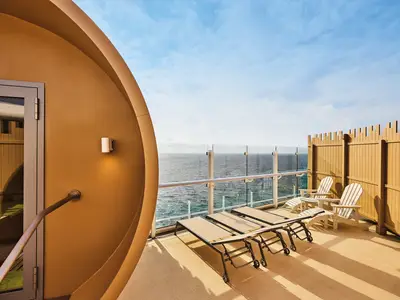 Panoramablick auf das Außendeck der Mein Schiff Relax mit einer braunen Fasssauna, hellen Sonnenliegen und zwei weißen Adirondack-Stühlen. Im Hintergrund erstreckt sich das weite blaue Meer unter einem klaren Himmel, umgeben von einer Glasbrüstung.