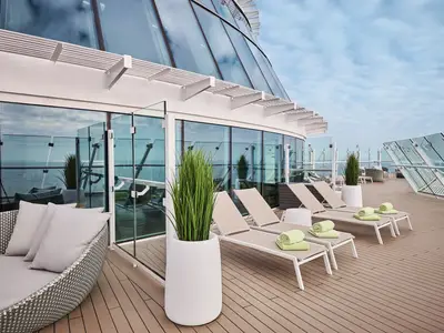 Blick auf das obere Sonnendeck der Mein Schiff Relax mit modernen Liegestühlen, einem Korbsessel und Glasgeländern. Im Hintergrund ist das offene Meer und der Himmel zu sehen. Grüne Handtücher liegen auf einigen Liegen bereit.