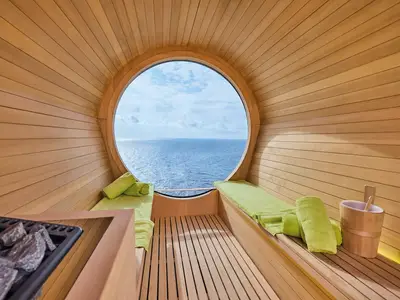 Gemütliche Holz-Fasssauna an Bord der Mein Schiff Relax. Ein grosses Rundfenster bietet Meerblick. Grüne Handtücher liegen auf den Holzbänken; links ist der Saunaofen zu sehen.