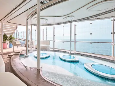 Heller Spa-Wellnessbereich auf der Mein Schiff Relax. Drei ovale Wasserbecken mit Wasserfall und dahinter Sitzbereiche auf dem Deck mit Meerblick.