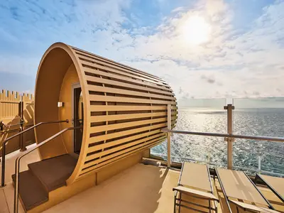 Moderne Fasssauna auf dem Sonnendeck der Mein Schiff Relax. Stufen zum Eingang. Glasreling, glitzerndes Meer und blauer Himmel mit Wolken. Liegestühle.