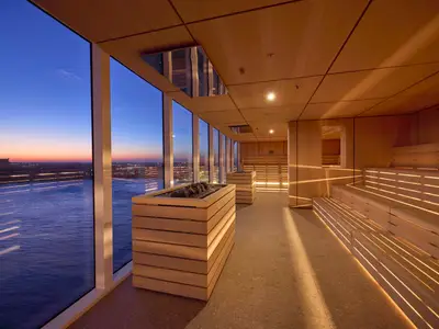 Moderne Horizontsauna an Bord der Mein Schiff Relax. Helle Holzsauna mit beleuchteten Bänken. Fensterblick auf Meer und beleuchtete Skyline bei Abenddämmerung. Zwei Steinbehälter, menschenleer.