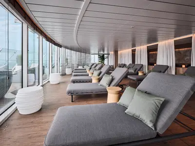 Innenansicht des Entspannungsbereichs an Bord der Mein Schiff Relax mit einer Reihe grauer Liegestühle und Kissen entlang einer geschwungenen Fensterfront. Im Hintergrund sind weitere Liegen und Vorhänge zu sehen, die eine ruhige Atmosphäre schaffen.