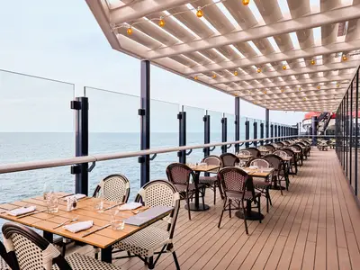 Outdoor Restaurantbereich an Bord der Mein Schiff Relax. Gedeckte Tische und Korbstuehle stehen unter einer Pergola mit Lichterketten, mit Blick auf das offene Meer.