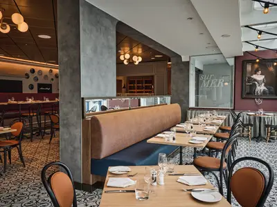 Das elegante sur Mer Bistrot auf der Mein Schiff Relax. Es sind Holztische, orangefarbene Stühle und bequeme Polsterbänke sichtbar. Eine Kunstinstallation schmückt die Wände.