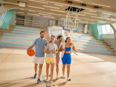 Vier lächelnde Freunde auf einem modernen Basketballfeld in der Sport-Arena an Bord der Mein Schiff Relax. Ein Mann hält einen Basketball, dahinter Sitzreihen und ein Korb.
