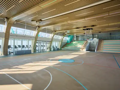 Helle Arena an Bord der Mein Schiff Relax mit einem Basketballfeld und hellblauen Markierungen. Links sind durch Fenster Fitnessgeräte und das Meer zu sehen. Rechts moderne, mehrfarbige Tribünen.