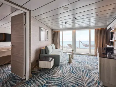 Innenansicht einer modernen Fernweh Suite an Bord der Mein Schiff Relax. Der Wohnbereich mit Sofa und Minibar führt zum Balkon mit Hängeliege und weitem Meerblick.