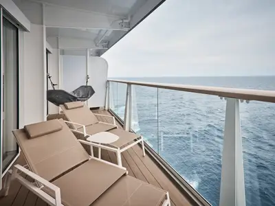 Geräumiger Balkon einer Suite an Bord der Mein Schiff Relax mit zwei beigen Liegestühlen, einem weißen Beistelltisch und einer grauen Hängematte. Im Hintergrund ist das weite blaue Meer zu sehen.