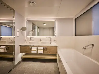 Elegantes Badezimmer einer Suite an Bord der Mein Schiff Relax mit Doppelwaschbecken, beleuchtetem Spiegel, modernen Holzmöbeln und einer Badewanne.