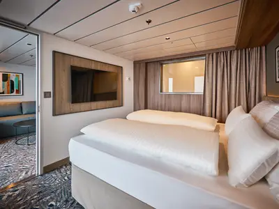 Schlafzimmer einer Weitblick Suite an Bord der Mein Schiff Relax. Weisses Doppelbett, Fenster mit Vorhang, Wandfernseher und Blick in den Wohnbereich mit Sofa.